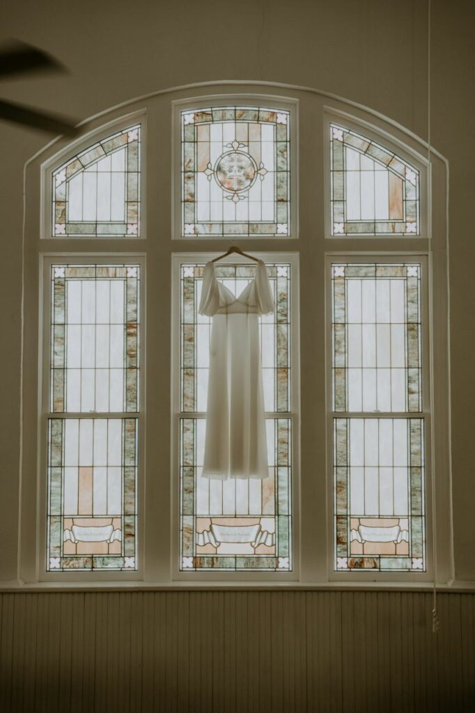 bride's wedding dress showcased in front of a stunning stained glass window at White Chapel & Harbor Hall.
